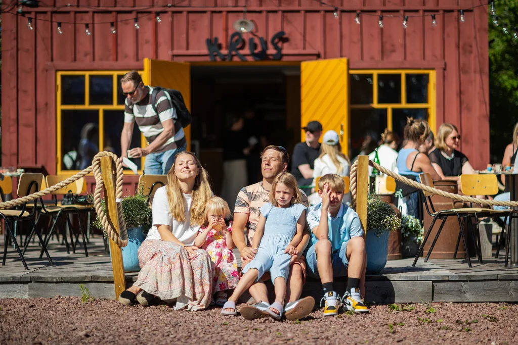 Familj sitter i solen framför rött hus med texten KRUS.