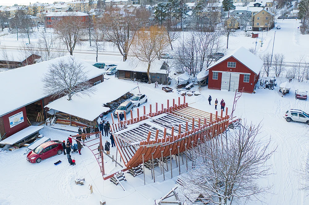 Flygfoto av människor som samlas runt en stor träbåtsram under konstruktion på en snöig gård, omgiven av röda byggnader, parkerade bilar och kala träd i ett vinterlandskap.